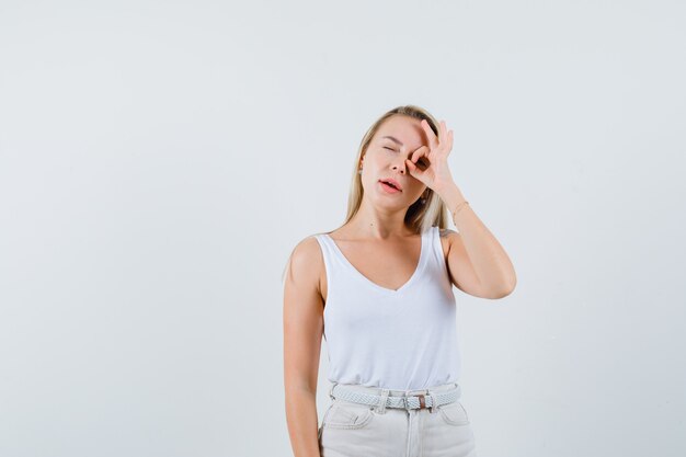 Señora rubia en camiseta, pantalones mostrando signo de ok en el ojo y mirando confiado