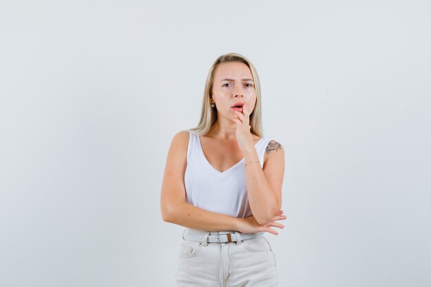 Señora rubia en camiseta, pantalones de pie en pose de pensamiento y mirando confundido