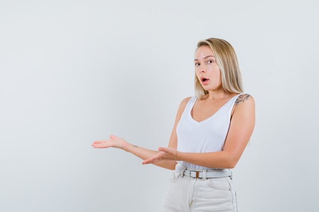 Señora rubia en camiseta, pantalones pretendiendo mostrar algo y mirando sorprendido, vista frontal.