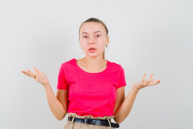 Señora rubia en camiseta rosa, pantalones mostrando un gesto de impotencia y mirando confundido, vista frontal.