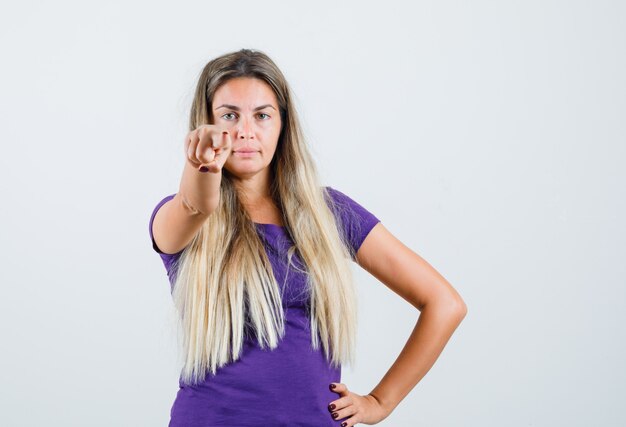 Señora rubia en camiseta violeta apuntando al frente y mirando confiada, vista frontal.