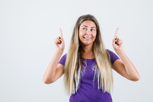 Señora rubia en camiseta violeta apuntando hacia arriba y mirando alegre, vista frontal.