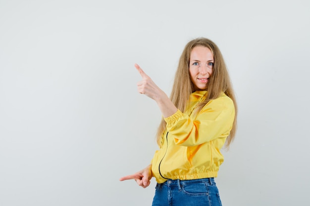 Señora rubia en chaqueta, jeans apuntando con el dedo hacia arriba y hacia abajo y mirando confiado,