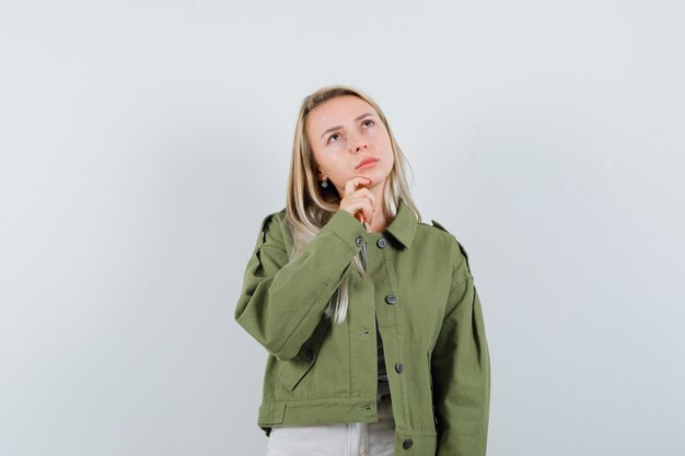 Señora rubia en chaqueta, pantalones sosteniendo la mano en la barbilla y mirando pensativa, vista frontal.