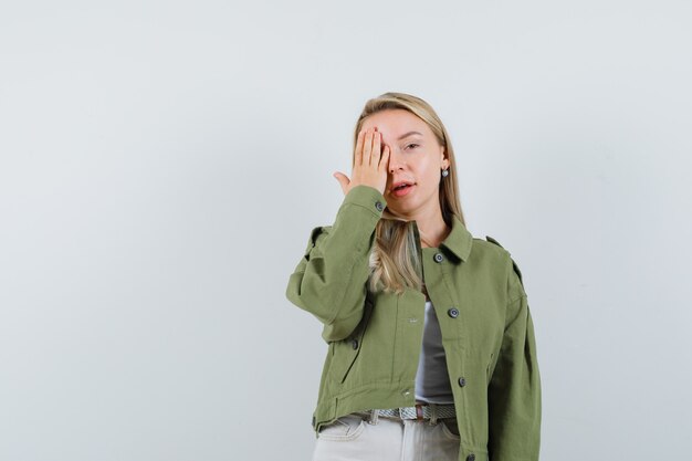 Señora rubia en chaqueta, pantalones sosteniendo la mano en el ojo y mirando positiva, vista frontal.