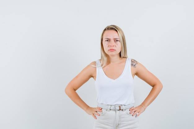 Señora rubia cogidos de la mano en la cintura en camiseta, pantalones y mirando rencorosa. vista frontal.