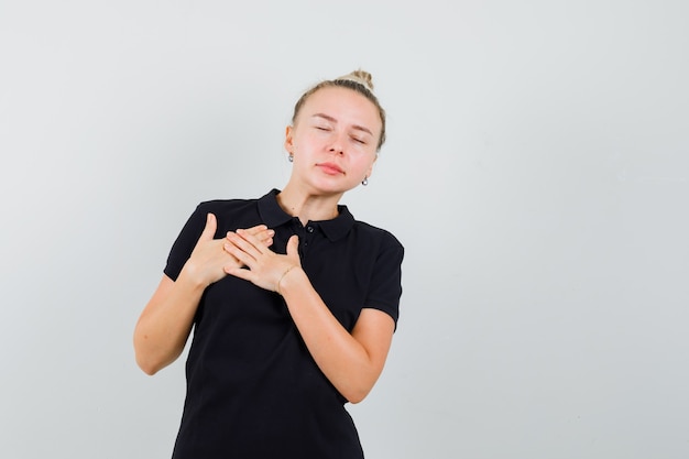 Foto gratuita señora rubia cogidos de la mano en el pecho en camiseta negra y mirando agradecido. vista frontal.