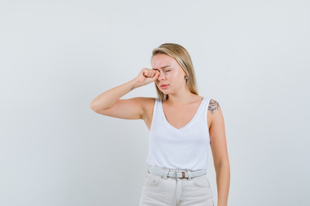 Señora rubia frotándose los ojos mientras llora en camiseta, pantalones y mirando ofendida. vista frontal.