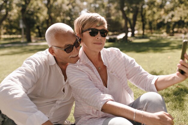 Señora rubia con gafas de sol, blusa con estilo a rayas y jeans. sentado en la hierba y haciendo selfie con hombre bigotudo con camisa blanca al aire libre.