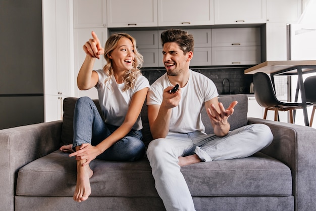 Señora rubia interesada ver la televisión. Retrato interior de una pareja sonriente sentada en un sofá acogedor.