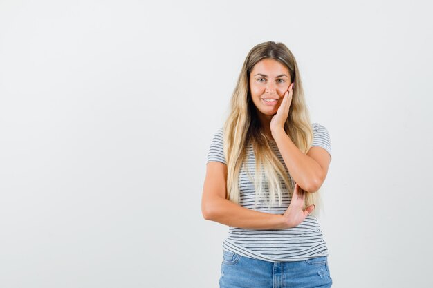 Señora rubia con la mano en la cara en camiseta y mirando alegre, vista frontal.