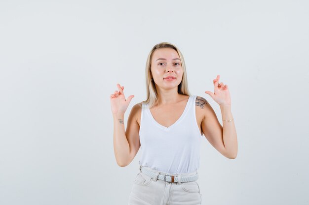 Señora rubia manteniendo los dedos cruzados en camiseta, pantalones y luciendo alegre