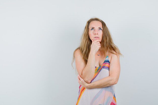 Señora rubia mirando hacia arriba con la mano en la barbilla en vestido de verano y mirando pensativo