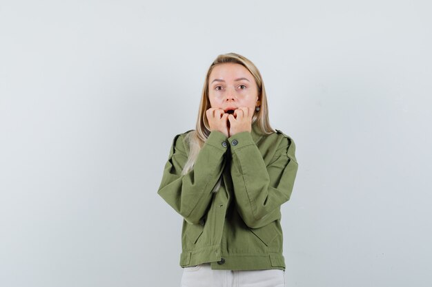 Señora rubia mordiendo las uñas emocionalmente en chaqueta, pantalones y mirando asustada. vista frontal.