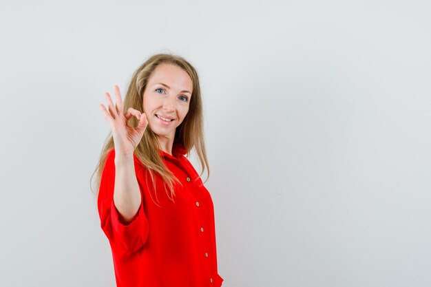 Señora rubia mostrando gesto ok en camisa roja y mirando feliz,