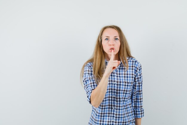 Señora rubia mostrando gesto de silencio en camisa y mirando con cuidado,