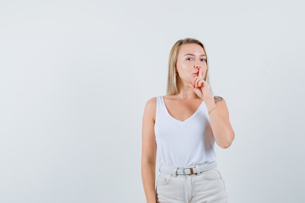 Señora rubia mostrando gesto de silencio en camiseta, pantalones y mirando confiado