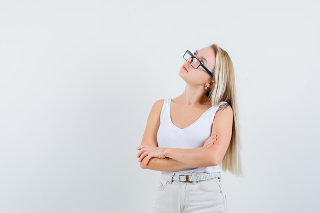 Señora rubia de pie con los brazos cruzados en camiseta, pantalones y mirando pensativa. vista frontal.