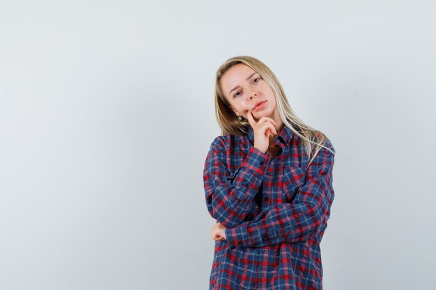 Señora rubia de pie en pose de pensamiento en camisa casual y mirando sensible, vista frontal.