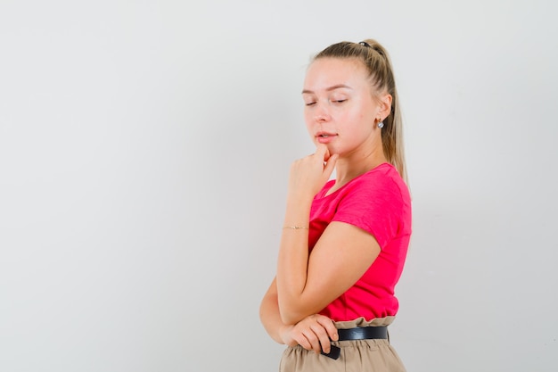 Señora rubia de pie en pose de pensamiento en camiseta rosa, pantalones y bonita.