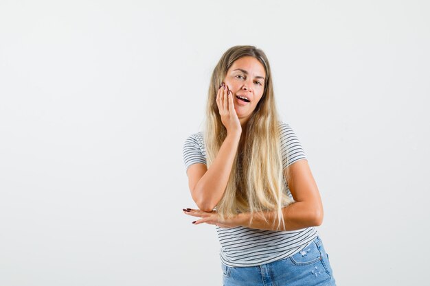 Señora rubia poniendo la mano en la cara en camiseta y mirando confiada. vista frontal.