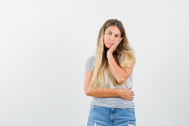 Señora rubia poniendo su mano en su barbilla en camiseta y mirando aburrida, vista frontal.