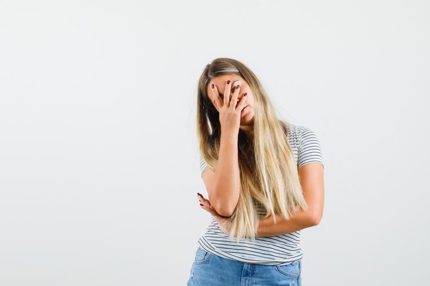 Señora rubia que cubre la cara con la mano en la camiseta y parece aburrida, vista frontal.