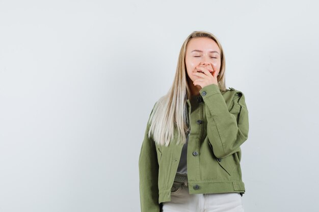 Señora rubia que lleva a cabo la mano en la boca en la chaqueta, los pantalones y parece contenta. vista frontal.