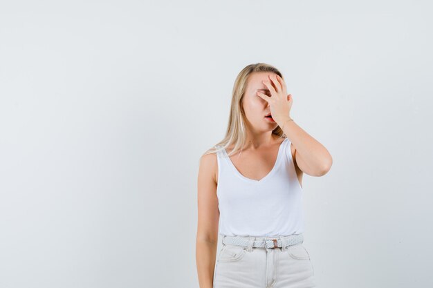 Señora rubia que lleva a cabo la mano en la cara en camiseta, pantalones y que parece angustiada. vista frontal.