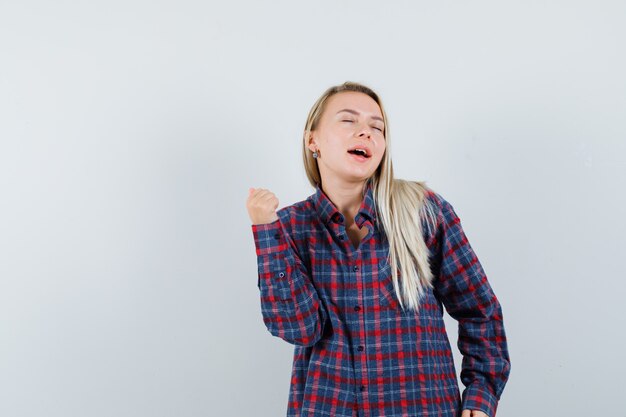 Señora rubia que muestra el gesto del ganador en camisa casual y mirando dichoso, vista frontal.