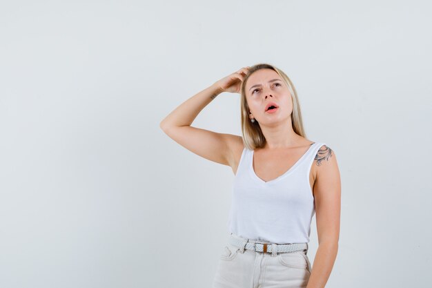 Señora rubia sosteniendo la mano en la cabeza en camiseta, pantalones y mirando pensativo, vista frontal.