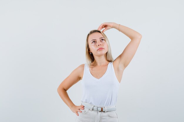 Señora rubia sosteniendo la mano en la cabeza en camiseta, pantalones y mirando pensativo