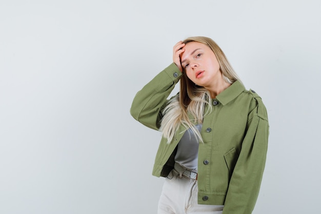 Señora rubia sosteniendo la mano en la cabeza en la chaqueta, pantalones y mirando pensativo, vista frontal.