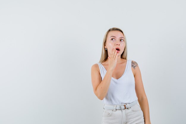 Señora rubia sosteniendo la mano cerca de la boca abierta en camiseta, pantalones y mirando sorprendido, vista frontal.