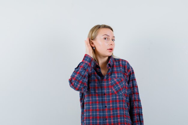 Señora rubia sosteniendo la mano detrás de la oreja en camisa casual y mirando curiosa. vista frontal.