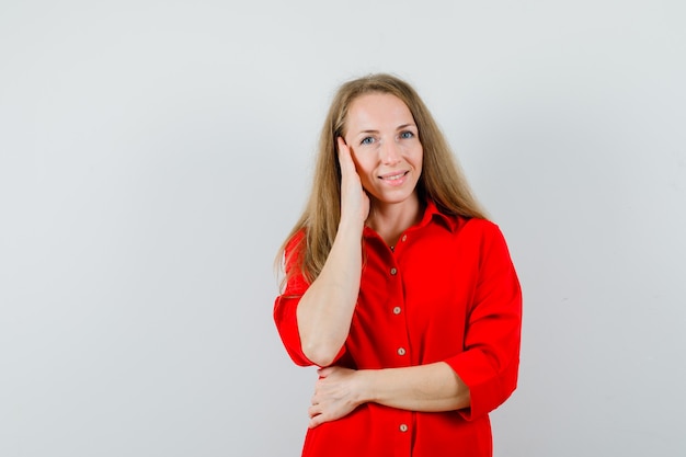 Señora rubia sosteniendo la mano en la mejilla con camisa roja y luciendo linda,