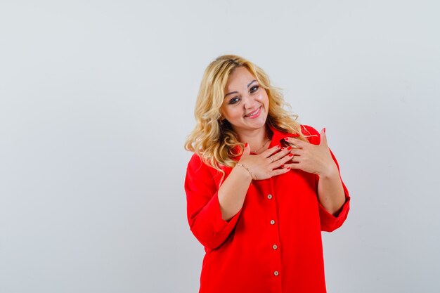 Señora rubia sosteniendo la mano sobre su pecho con camisa roja y mirando alegre espacio para texto