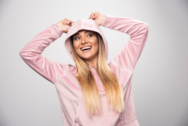 Señora rubia en sudadera rosa haciendo poses alegres y positivas usando una sudadera con capucha en la cabeza.