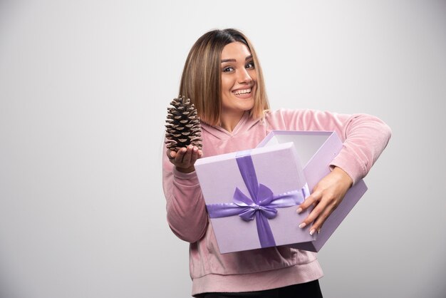 Señora rubia con sudadera rosa saca un cono de roble de la caja de regalo y se siente feliz.