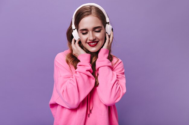 Señora rubia en sudadera rosa sonriendo y escuchando música en auriculares