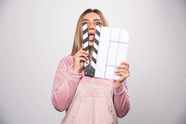 Señora rubia en sudadera rosa sosteniendo un tablero de azote en blanco y pone una parte de su cuerpo a través de él.