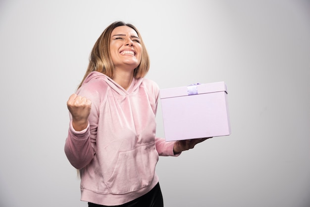 Señora rubia en sudadera rosa tiene una caja de regalo y se ve muy feliz.