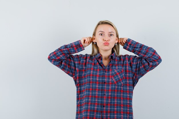 Señora rubia tirando de sus orejas, haciendo pucheros con los labios en una camisa casual y luciendo divertida, vista frontal.