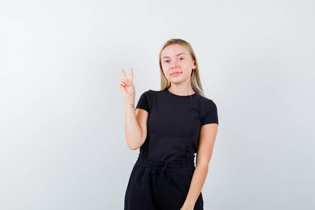 Señora rubia vestida de negro que muestra el signo V y que parece segura, vista frontal.