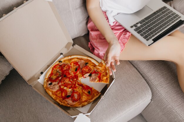 Señora sentada en el sofá con laptop y comida rápida. Freelancer mujer caucásica comiendo pizza durante el trabajo con la computadora.