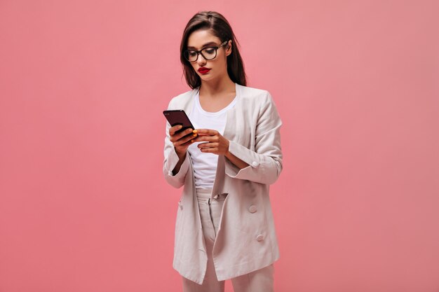 Señora seria en anteojos y traje charlando por teléfono. Hermosa morena con labios rojos en traje beige sostiene smartphone sobre fondo aislado.