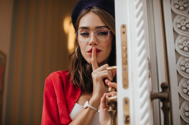 Señora sexy con cabello castaño, lápiz labial rojo en boina, camisa, accesorios elegantes posando y sosteniendo el dedo índice en los labios, mirando al frente y abriendo puertas blancas