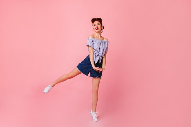 Señora soñadora en pantalones cortos de mezclilla bailando con una sonrisa. Hermosa chica pinup saltando sobre el espacio rosa.