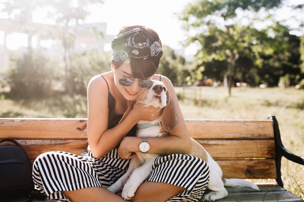 Foto gratuita señora sonriente bronceada en elegante reloj de pulsera abrazando perro beagle durante el descanso en el parque por la mañana