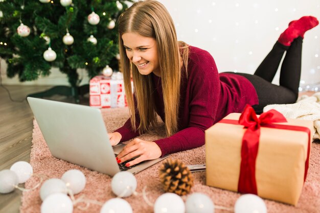 Señora sonriente con la computadora portátil cerca de cajas de regalo, gancho, luces de hadas y árbol de Navidad